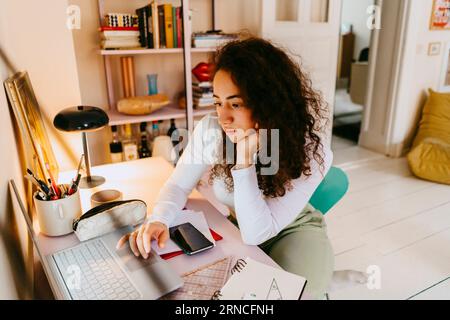 Young woman with curly hair using laptop while studying at home Stock Photo