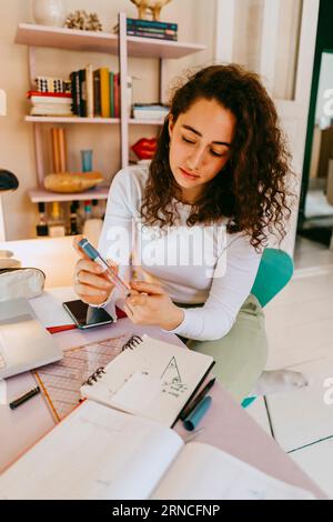 Young woman with curly hair using glucometer while doing homework at home Stock Photo