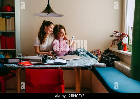 Happy young female friends sharing smart phone while sitting at home Stock Photo