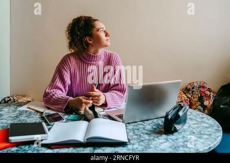 Thoughtful young woman looking away while sitting with laptop and books at table Stock Photo