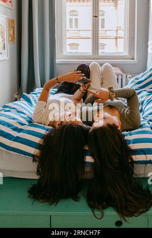 Young female friends using smart phone while lying on bed at home Stock Photo