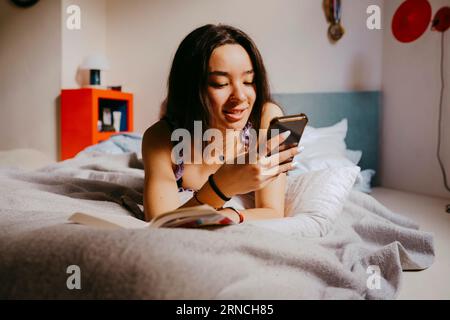 Smiling young woman using smart phone while lying on bed at home Stock Photo