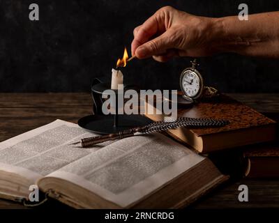 A female hand lights a candle on a table with old books, a quill pen and an antique pocket watch Stock Photo