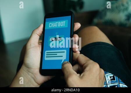 a young man, in casual wear, is using his smartphone, that reads the text chatbot in its screen, while is sitting comfortably in a chair Stock Photo
