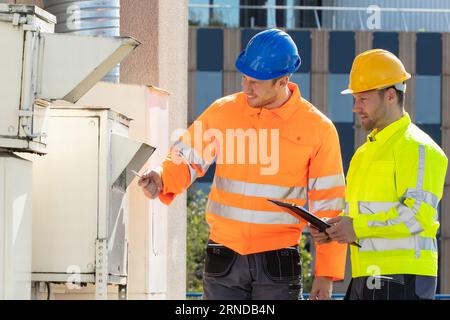 Two Electricians Men Wearing Safety Jackets Checking Air Conditioning Unit On Building Rooftop Stock Photo