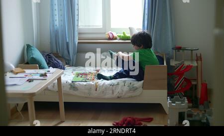 Candid little boy trying to put socks sitting in bed inside bedroom ...