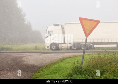 Yield sign on the side of a rural road, with white semi trailer truck passing by on highway at speed on a rainy, foggy day with low visibility. Stock Photo