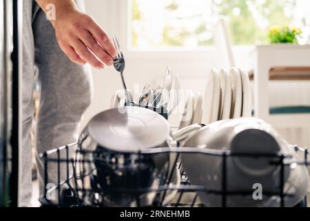 Dish washer machine in kitchen. Man loading dishwasher. Washing plates. Fork in hand. Full of cutlery. Clean or dirty. Pot and tableware. Stock Photo