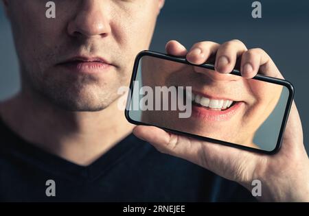 Fake social media smile or filter. Sad face and happy in phone screen. Mental health, depression online and technology concept. Man with anxiety. Stock Photo