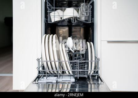 Dish washer machine. Washing plates and cutlery in basket. Open dishwasher in kitchen, front view. Full load of clean utensils and tableware. Stock Photo