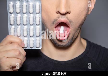 sick man with wide open mouth has capsule on his tongue and holds blister up next to his face  while taking pill against pain Stock Photo