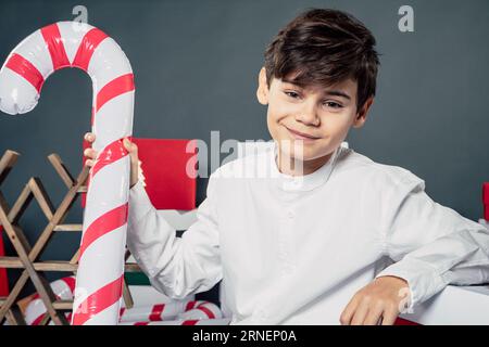 young teen boy holding inflatable candy cane in hand  and white shirt sit on floor with xmas presents in background and smiling into camera  and be ha Stock Photo