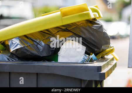 full garbage trash can with ffp2 face mask standing at sidewalk waiting to get empty the recyclable plastic dump out of the black yellow container and Stock Photo