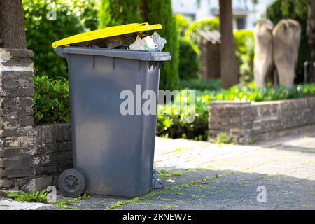 full garbage trash can standing at sidewalk next to street waiting to get collected and empty the recyclable plastic dump out of the black yellow cont Stock Photo