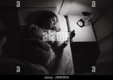A pregnant woman lies in bed and uses a smartphone. Insomnia. Stock Photo
