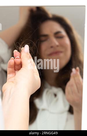 Young girl with a disgusted look who, looking at herself in the mirror, observes a lock of hair between her fingers. Trichotillomania disorder, a comp Stock Photo