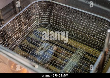 Empty industrial deep fryer filled with hot oil. Visible metal mesh or basket for fries or other food for frying. Stainless steel used around. Stock Photo
