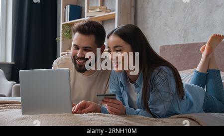 Caucasian couple family lying together on bed use credit bank card enter code in laptop computer banking app online payment smiling woman and man Stock Photo