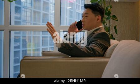Asian happy businessman chinese japanese man calling mobile phone sit in office modern company smiling ethnic middle-aged entrepreneur employee worker Stock Photo