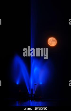 A blue super moon rises beside a water fountain coloured blue over Little Lake in Peterborough, Ontario. Stock Photo