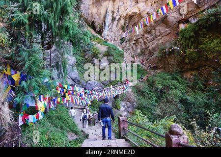 Hikers walk toward colorful prayer flags over the trail as they approach the final ascent to the Tiger's Nest Monastery (Taktsang) near Paro, Bhutan Stock Photo