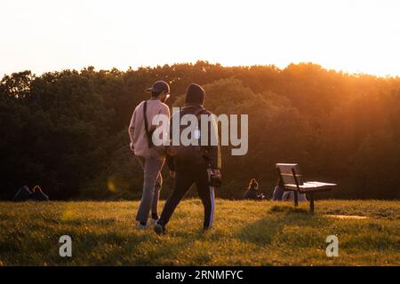 Young Men walking towards wooden park bench during golden hour sunset. People, relationships, enjoying Stock Photo