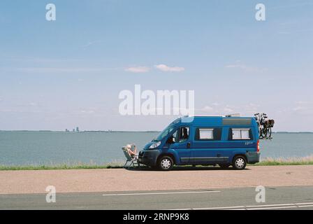 A senior woman on holiday sits in a beach chair near a camper van next to sea, lounging Stock Photo