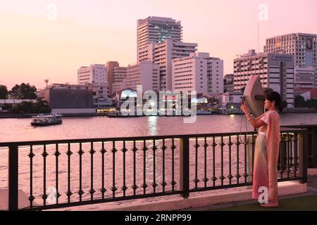 Thai woman in traditional costume as part of a river tour takes a photo at sunset near the Chao Phraya river Stock Photo
