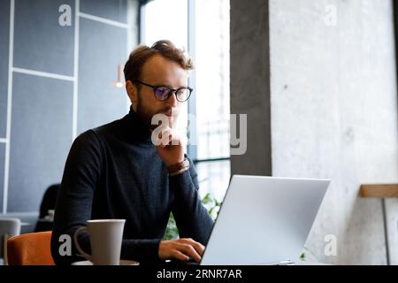 A young red-haired guy, a programmer or entrepreneur in glasses, works in a stylish cafe behind a laptop. Freelancer works remotely. Online Stock Photo