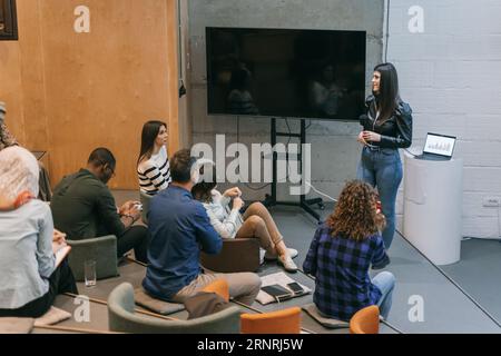 Young and beautiful business woman giving a lecture to a diverse group of employees in a small lecture hall and discussing with them Stock Photo