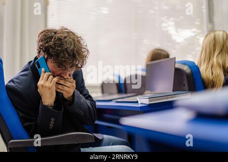 Male student talking on his phone during class instead of paying attention to the professor Stock Photo