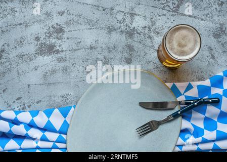 Oktoberfest table setting with blue checkered tablecloth, cutlery, empty plate and mug of beer on grey concrete background Stock Photo