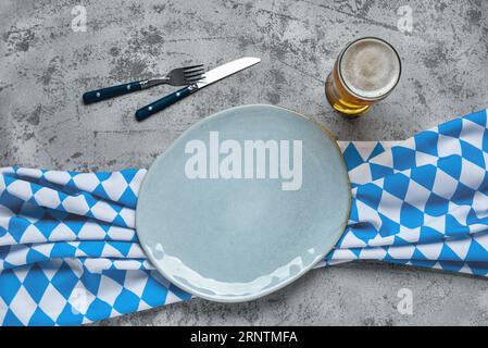 Oktoberfest table setting with blue checkered tablecloth, cutlery, empty plate and mug of beer on grey concrete background Stock Photo