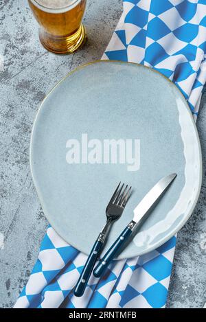 Oktoberfest table setting with blue checkered tablecloth, cutlery, empty plate and mug of beer on grey concrete background Stock Photo