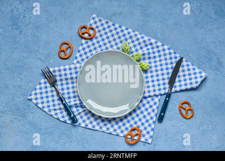 Oktoberfest table setting with empty plate, cutlery, pretzel, hops and checkered napkin on blue background Stock Photo