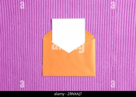 Orange envelope with blank card on purple knitted fabric background. Birthday, invitation, holiday concept. Top view, flat lay, mockup. Stock Photo