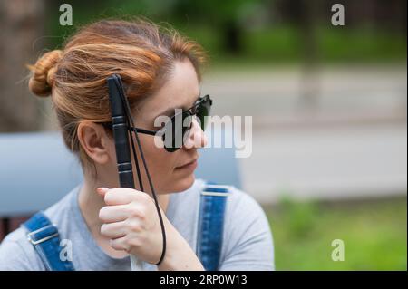Blind red-haired woman sitting outdoors leaning on a cane.  Stock Photo