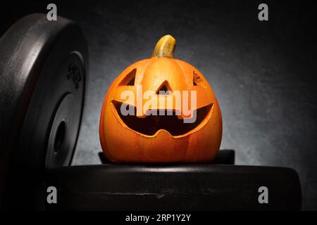 Halloween pumpkin decoration and dumbbell barbell weight plates. Healthy fitness autumn fall spooky composition. Gym workout, sport training concept. Stock Photo