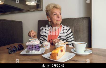 Attractive, joyful older woman drinking coffee while sitting at a cafe. Portrait of mature woman in a cafeteria. Colorful cakes and coffee on the Stock Photo