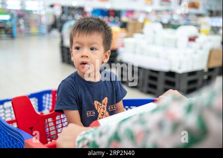 Two year old adorable multiracial toddler in a blue t-shirt sitting in a shopping trolley with groceries, in grocery store Stock Photo