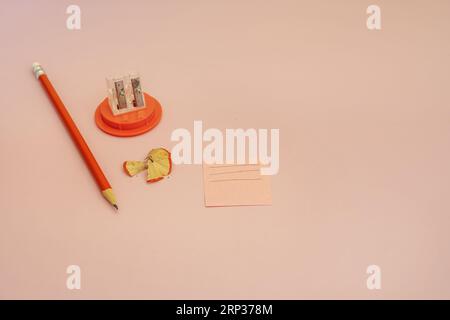 A sharpened orange pencil with a small eraser on one end next to a pencil sharpener, a note and wooden peels Stock Photo