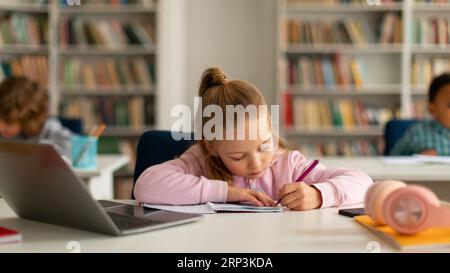 Focused caucasian pupil girl writing in copybook while using laptop computer, sitting at desk at elementary school Stock Photo
