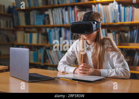 Girl student using modern tech devices in the library, a laptop and VR headset. Concepts of virtual reality experience in education. Stock Photo