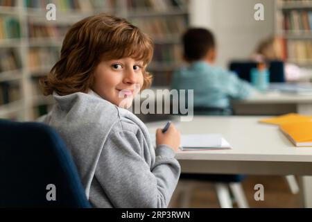 Smart male elementary school pupil, sitting at desk in classroom, turning back and smiling at camera during lesson Stock Photo