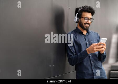 Millennial eastern man standing by black wall, using phone, headphones Stock Photo