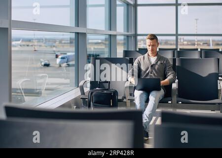 Traveling by airplane. Concentrated man working on laptop while waiting for flight. Solo traveler with suitcase at airport. Stock Photo
