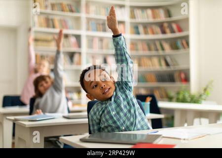 Diverse elementary school students raising hands to answer teacher questions, sitting at desks in classroom interior Stock Photo