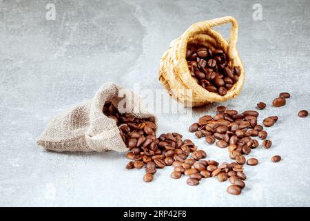 A pile of coffee beans spilled out of a cane basket and burlap bag on a gray abstract background. Stock Photo