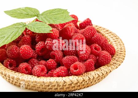 Berries of ripe raspberries in a wicker basket on a white background. On top of a pile of berries lies a green leaf of a raspberry bush Stock Photo
