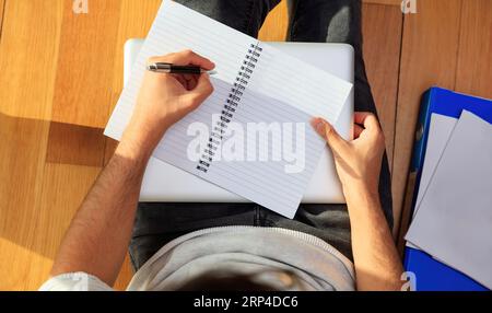 Young man is sitting on the wooden floor and making note on empty notebook paper. Workspace at home. Top view, copy space Stock Photo